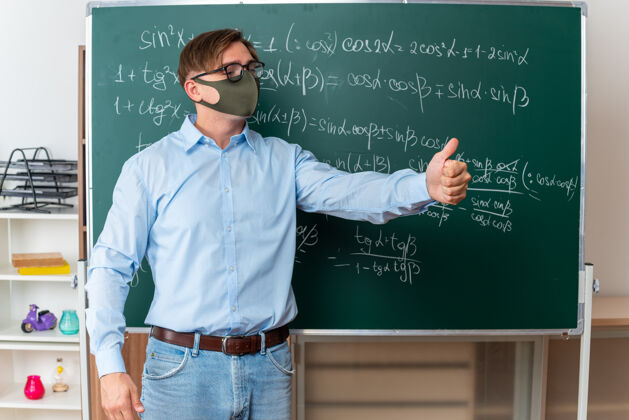 黑板戴着护目镜 戴着口罩的年轻男教师在教室里讲解数学公式 站在黑板旁竖起大拇指课站面部