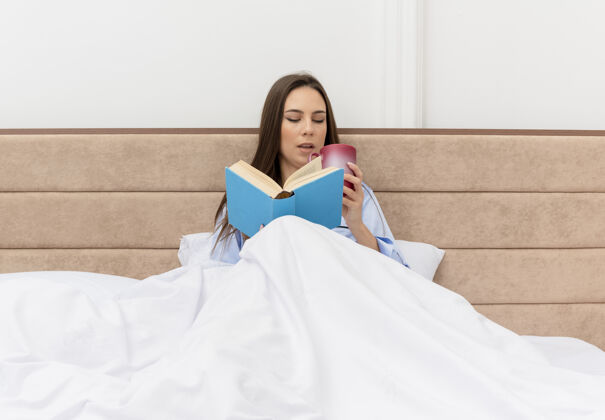 周末穿着蓝色睡衣的年轻美女躺在床上 拿着书和咖啡 微笑着看书 在卧室里享受周末床灯光美丽
