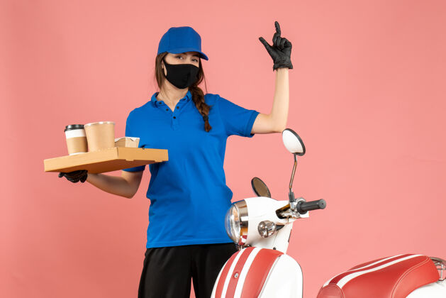 前面正面图微笑的快递员女孩戴着医用口罩手套站在摩托车旁边拿着咖啡小蛋糕在粉彩桃色背景上抱小咖啡
