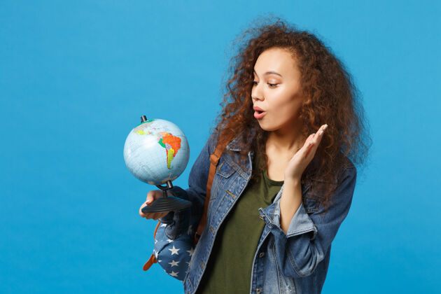 复制空间年轻的非洲裔美国女孩 穿着牛仔服 背着背包 把地球孤立地放在蓝色的墙上旅程地球旅游