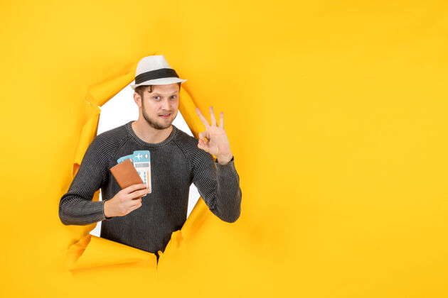 制造一个自信的年轻人戴着帽子 拿着外国护照和机票 在撕破的黄色墙上做着戴眼镜的手势帽子手势护照