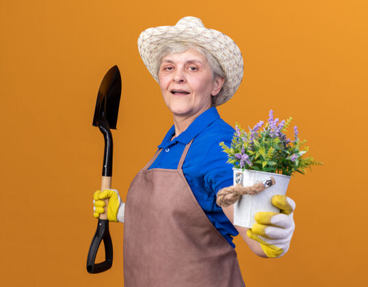 花园自信的上了年纪的女园丁戴着园艺帽和手套站在一边拿着铁锹和花盆侧着站着戴着