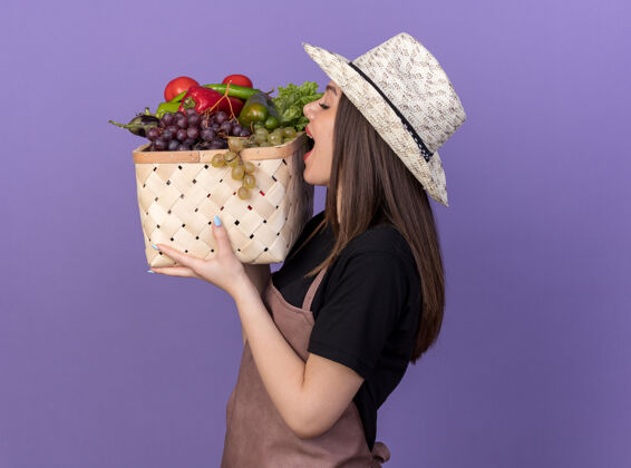 抱高兴的是漂亮的白人女园丁戴着园艺帽拿着并假装咬菜篮子花园篮子紫色
