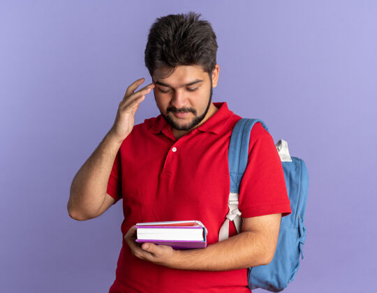 姿势年轻的留着胡子的学生 穿着红色马球衫 背着书包 看着书 微笑着 自信 快乐 积极地站在蓝色的墙上姿势正面站