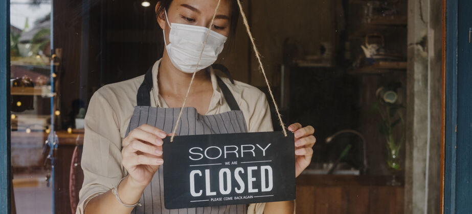 停止年轻的亚洲女孩戴着口罩 在冠状病毒封锁检疫后 在玻璃门咖啡馆的一个标志由开放变为关闭围裙餐厅年轻人
