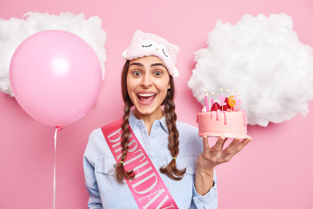 心情扎着两条辫子的女孩高兴地接受了祝贺 手里拿着美味的草莓蛋糕 充气的氦气球孤立在粉红色上睡眠面具乐观欢乐