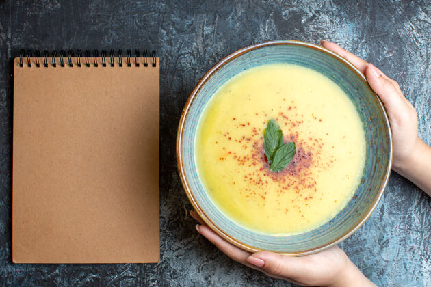 美食上图：手拿着一个蓝色的锅 里面有美味的汤和蓝色背景上的螺旋笔记本蔬菜汤碗汤