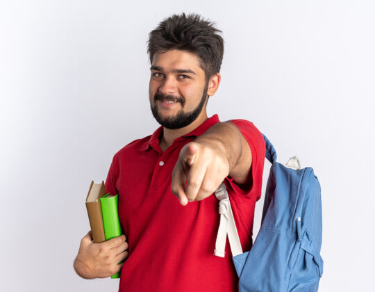 笔记本年轻的留着胡子的学生 穿着红色马球衫 背着背包 拿着笔记本 用食指指着 站在白色的墙上 快乐而积极地微笑着人指向手指