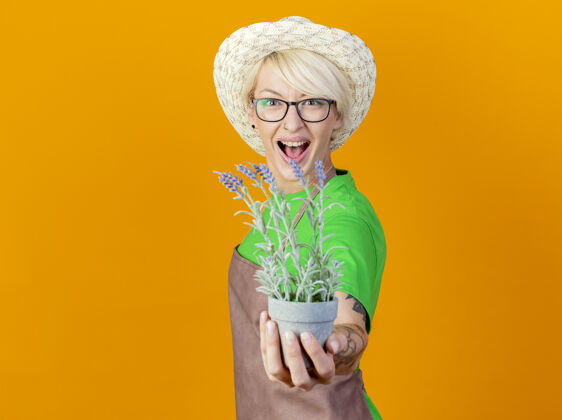 站一位年轻的园丁 围裙短发 戴着帽子 站在橙色的背景下 脸上挂着盆栽植物的微笑锅头发微笑