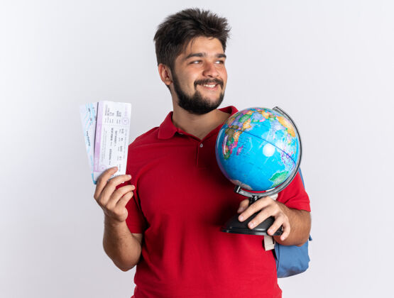 胡须年轻的留着胡子的学生 穿着红色马球衫 背着背包 手里拿着机票 站在白墙上开心地微笑着看着地球地球仪姿势马球