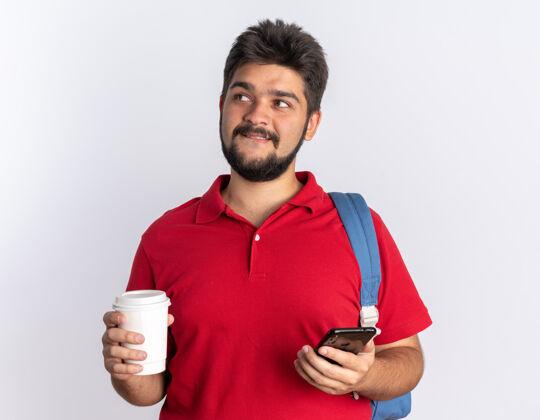 背包年轻的留着胡子的学生 穿着红色马球衫 背着背包 手里拿着智能手机和咖啡杯 站在白色的墙上 脸上带着狡黠的微笑手机年轻微笑