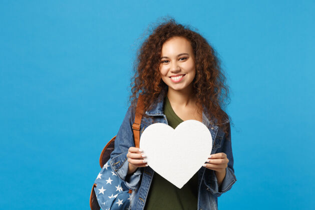 青少年年轻的非洲裔美国女孩十几岁的学生在牛仔服装 背包举行的心隔离在蓝色的墙上女孩空白空