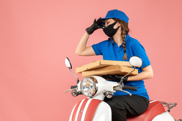 摩托车俯视图：戴着医用面罩和手套的女快递员坐在滑板车上传递订单 在柔和的桃色背景下做出难闻的手势头盔桃坏