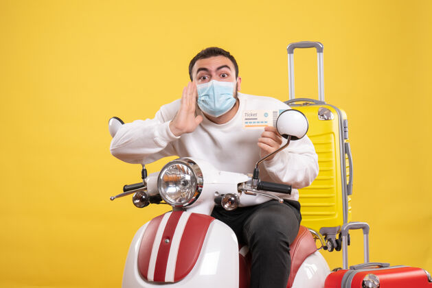 人旅行概念的俯视图 戴着医用面罩的年轻忧心忡忡的家伙坐在摩托车上 背着黄色手提箱 手里拿着车票头盔运动持有