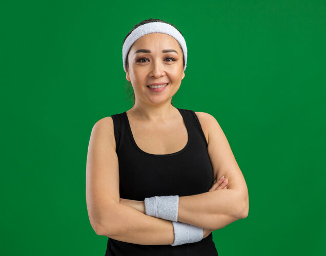 年轻戴着头巾和臂章 脸上挂着微笑 双臂交叉的年轻健身女士站在绿色的墙上头带十字架健康