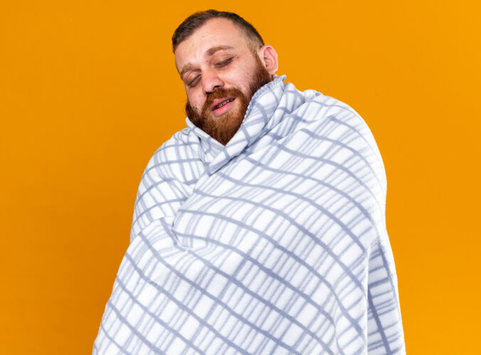 站不健康的胡子男人裹着毯子 感觉不舒服 站在橙色的墙上闭着眼睛 忍受着寒冷病痛苦冷