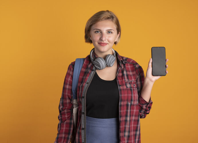 橙色高兴的年轻斯拉夫女学生戴着耳机背着背包拿着电话电话复制女孩