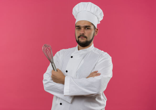 厨师自信的年轻男性厨师 身着厨师制服 站在粉红色的墙上 保持封闭的姿势 拿着威士忌立场封闭持有