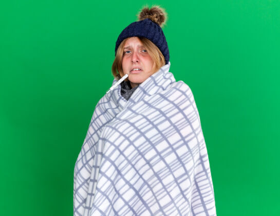 站立不健康的年轻女子裹着毯子戴着帽子用体温计测量体温患流感发烧站在绿色的墙上包裹年轻人毯子