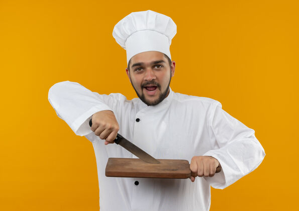 板印象深刻的年轻男厨师在厨师制服持刀和砧板隔离在橙色的墙上印象制服男性