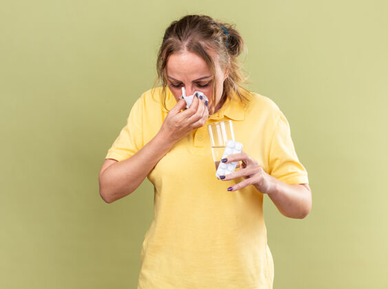 药片不健康的穿黄衬衫的女人 拿着一杯水和药片 站在绿色的墙壁上 流鼻涕 打喷嚏 打喷嚏跑步鼻子女人