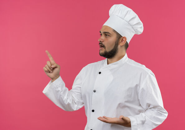 空穿着厨师制服的年轻男厨师 空手看着并指着粉色墙壁上隔离的一面 并留有复印空间厨师烹饪男性