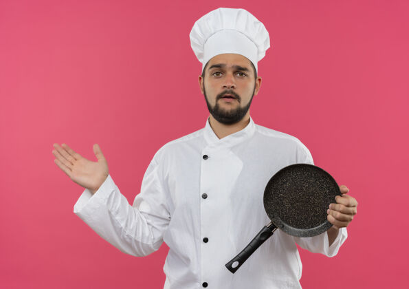 烹饪困惑的年轻男厨师身着厨师制服 手里拿着煎锅 在粉红色的墙上孤立着一只空手平底锅空显示