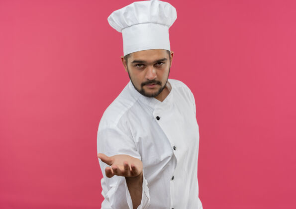 伸展自信的年轻男厨师穿着厨师制服 伸出手 在粉色的墙上与复制空间隔离厨师年轻手