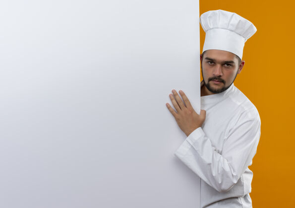 手自信的年轻男厨师身着厨师制服站在身后 将手放在白色的墙上 橙色的墙上有复制空间男性厨师信心