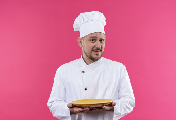 盘子自信的年轻帅哥厨师身着厨师制服 把盘子孤立在粉红色的墙上厨师制服年轻