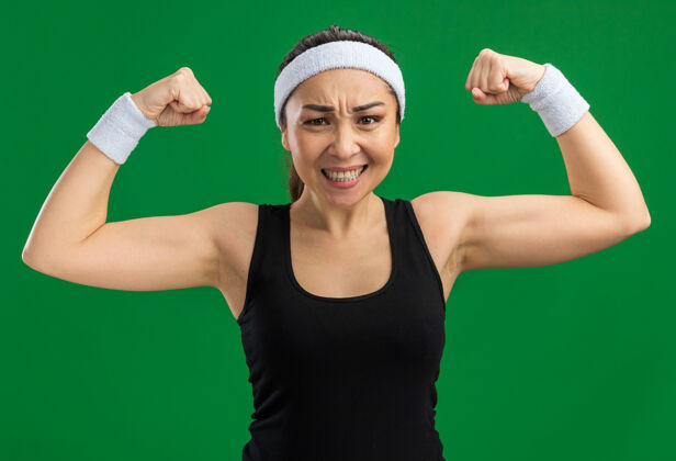 健康戴着头带和臂章的年轻健身女士绷紧了 自信地举起拳头 站在绿色的墙上 展现出力量和力量头带女人力量
