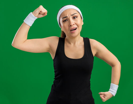 站立戴着头带和臂章的年轻健身女士绷紧了 自信地举起拳头 站在绿色的墙上 展现出力量和力量展示紧张女人