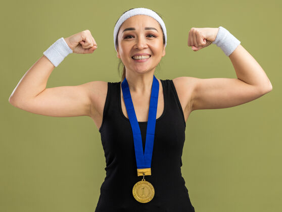 站立戴着头带和袖带 脖子上戴着金牌的年轻健身女士微笑着自信地举起拳头 展示着站在绿墙上的力量和力量自信头带健身