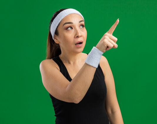 手指戴着头带和臂章的年轻健身女士一边看一边好奇地用食指指着绿色墙上站着的东西指点年轻女人