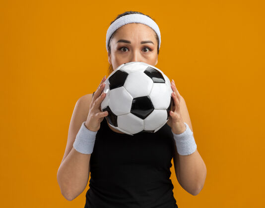 前面戴着头带和臂章的年轻健身女士 脸前拿着足球 站在橘色的墙上 她很担心年轻担心头带