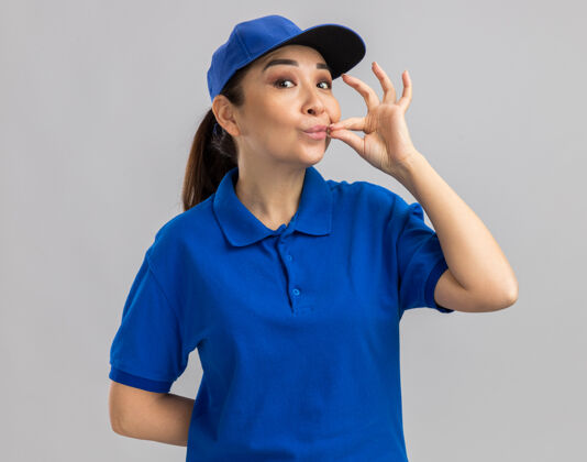 女人身穿蓝色制服 头戴鸭舌帽的年轻女送货员做着沉默的手势 就像站在白墙上的拉链合上了嘴关闭使手势