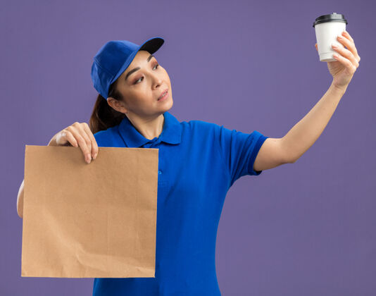 阴谋身穿蓝色制服 头戴鸭舌帽的年轻女送货员拿着纸包和纸杯 站在紫色的墙上 神情严肃地看着杯子制服交货帽子