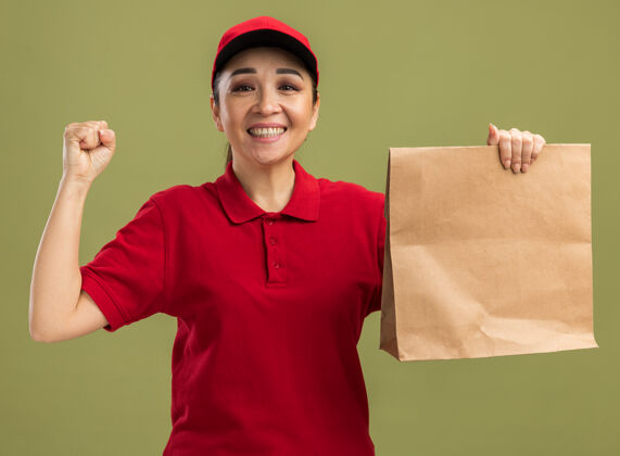 拳头快乐而兴奋的年轻送货员身穿红色制服 戴着帽子 手里拿着纸袋 脸上挂着微笑 握紧拳头站在绿色的墙上制服站着女人