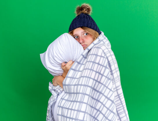 流感不健康的年轻女子裹着毛毯戴着帽子抱着枕头忍受着感冒和流感的折磨站在绿色的墙上毯子生病女人