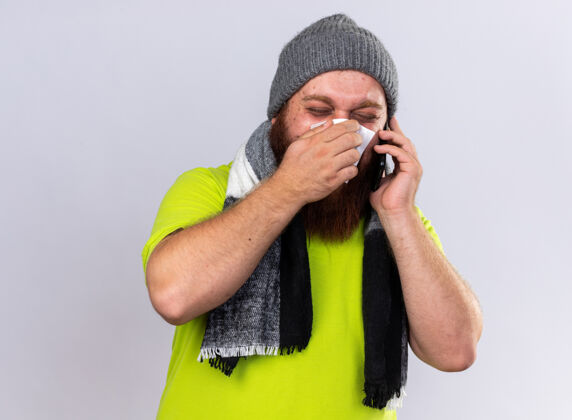 手机戴着帽子 围着暖和的围巾 留着不健康的胡须 脖子上戴着暖和的围巾 感觉很可怕 患流感 打手机 流鼻涕 站在白墙上打喷嚏打喷嚏感觉鼻子
