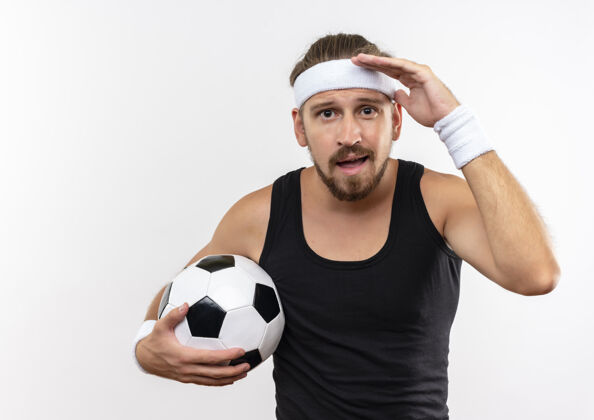 手令人印象深刻的年轻英俊的运动男子戴着头带和腕带拿着足球 把手放在额头附近隔离在白色的墙上运动男人近