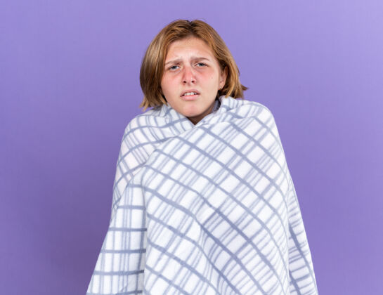 强壮不健康的年轻女子裹着温暖的毯子 感觉不舒服 患流感 发烧 头痛 站在紫色的墙上温暖女人生病
