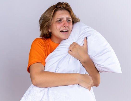 感觉不健康的年轻女子穿着橘色t恤 抱着枕头 站在白色的墙壁上感觉不舒服 患流感站着生病抱着