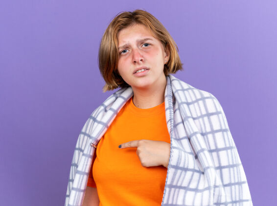 测量不健康的年轻女子裹着温暖的毯子 感觉不舒服 患流感 发烧 用温度计测量体温 站在紫色的墙上 看起来很担心包裹流感年轻