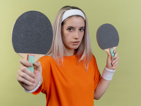 运动自信的年轻白种人运动女孩戴着背带戴着头带和手环拿着乒乓球拍乒乓球磨损背带