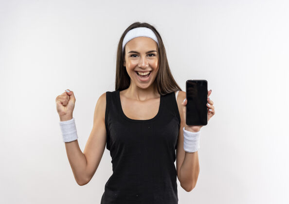 拳头快乐漂亮的运动女孩戴着头带和腕带手持手机 举起拳头隔离在白墙上握着穿手机