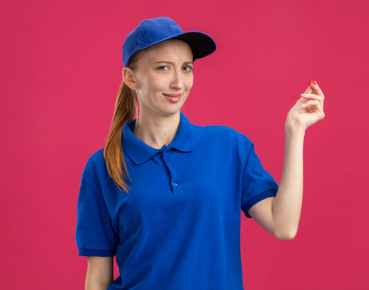 手指穿着蓝色制服 戴着帽子 微笑着挣钱的年轻送货女孩站在粉红色的墙上 用手指搓着制服送货钱