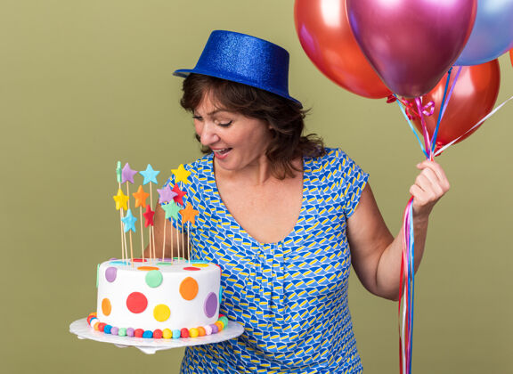帽子快乐的中年妇女戴着派对帽 手里拿着一堆五颜六色的气球 手里拿着生日蛋糕 看着它微笑着站在绿色的墙上庆祝生日派对中间束五颜六色