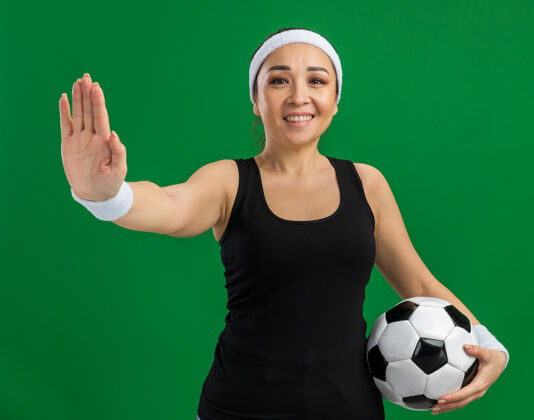 女人戴着头巾的年轻健身女 面带微笑地拿着足球 站在绿色的墙上做着停下来的手势开站年轻