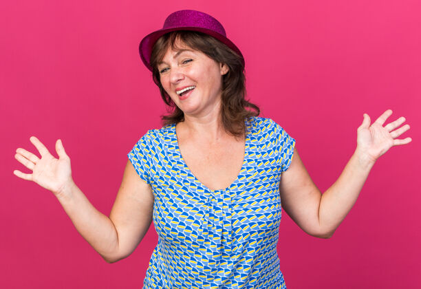 庆祝戴着派对帽的中年妇女高举双臂欢笑着站在粉色的墙上庆祝生日派对站立帽子女人